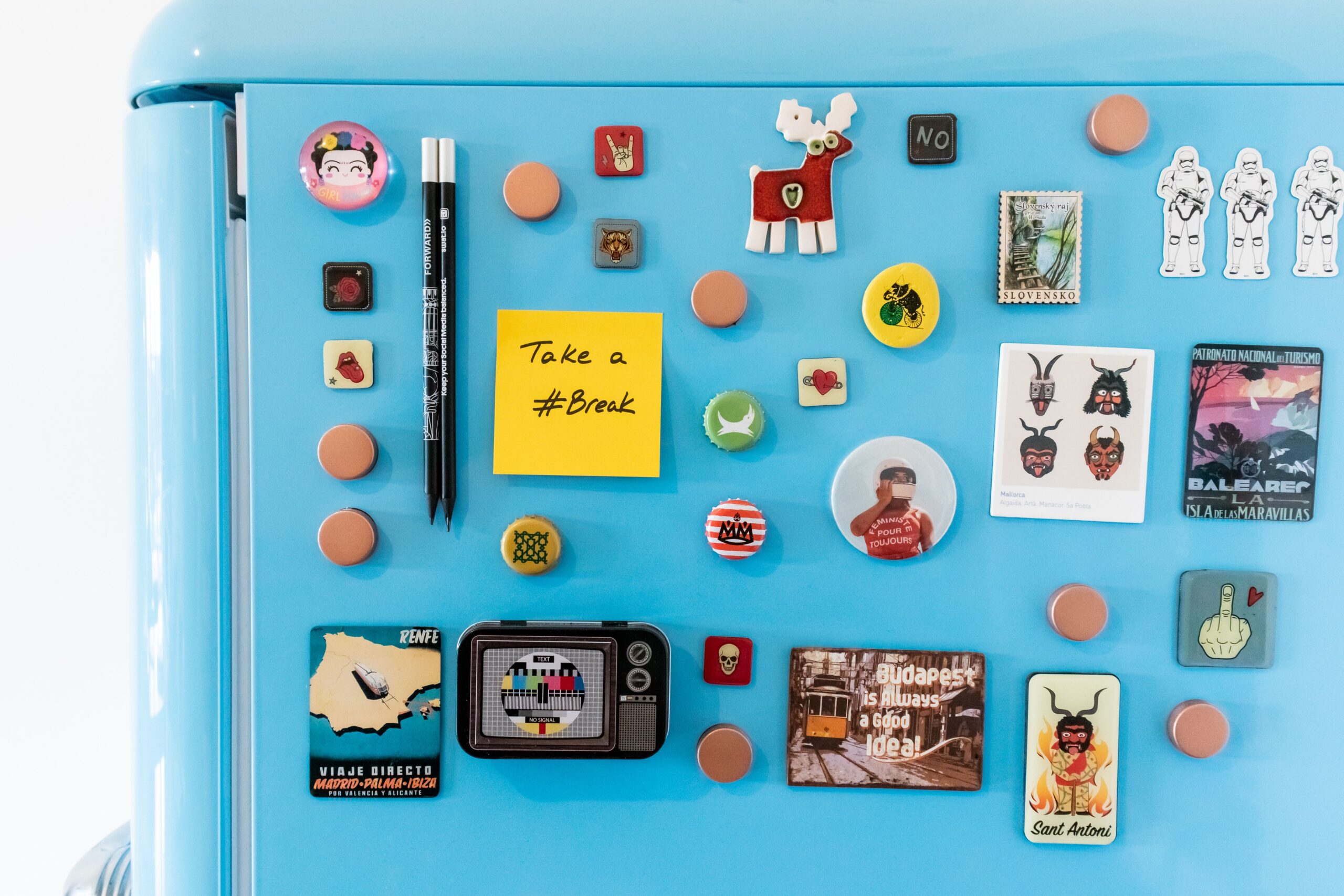 A vibrant collection of magnets on a blue fridge, featuring souvenirs and a sticky note reading 'Take a #Break'.